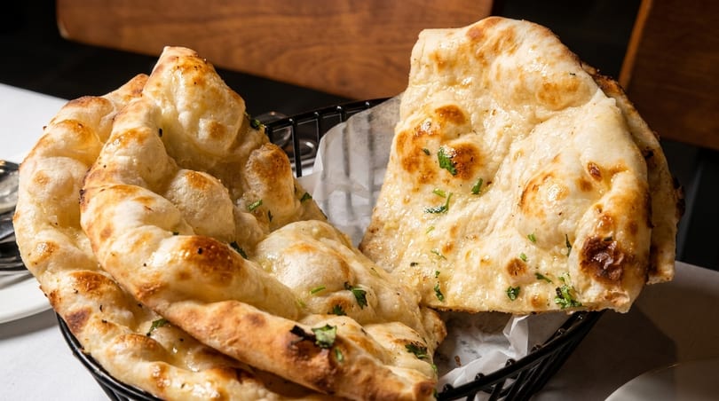 Golden-brown naan bread, garnished with fresh herbs, in a black wire basket on a white tablecloth.