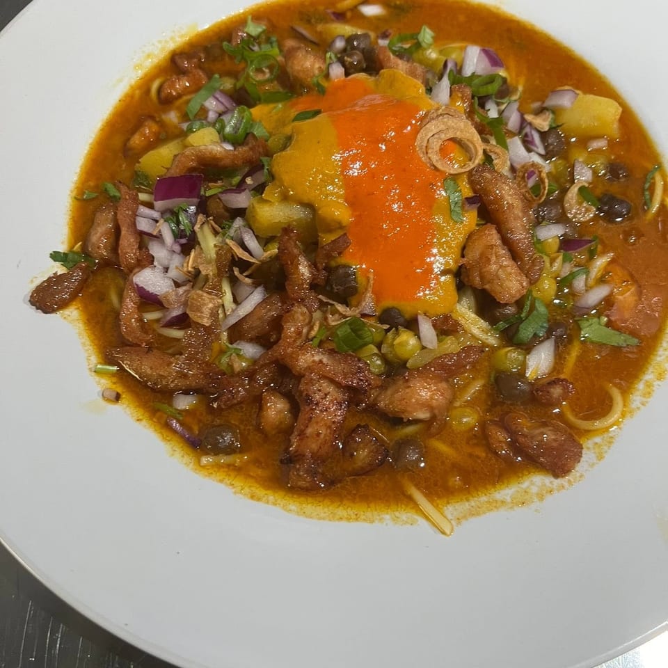 A vibrant bowl of curry noodles with chicken, red onions, green peas, and a dollop of orange sauce.
