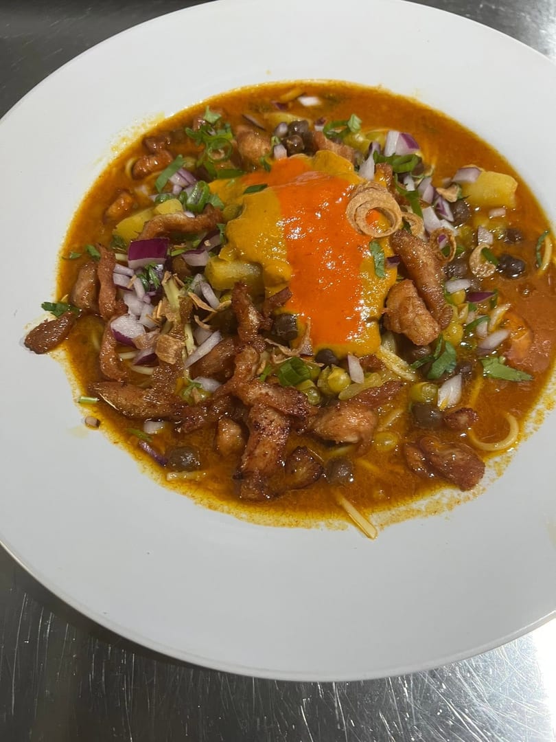 A vibrant bowl of curry noodles with chicken, red onions, green peas, and a dollop of orange sauce.