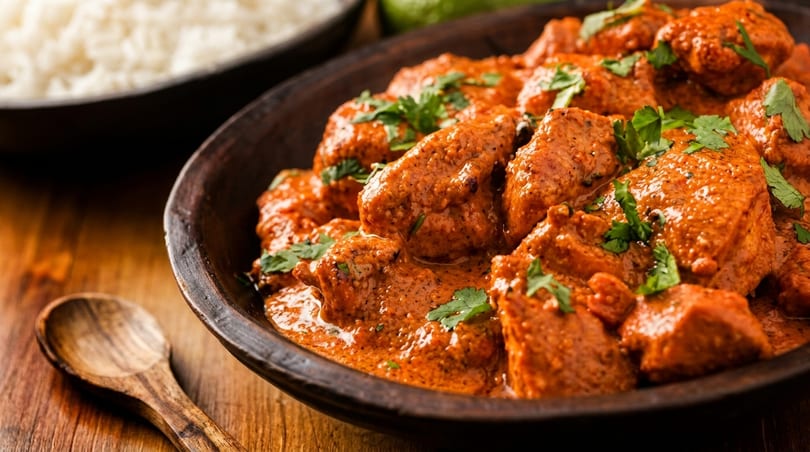 Close-up of chicken tikka masala in a dark bowl with rice and a wooden spoon.